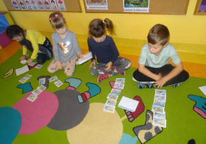 Czwórka dzieci siedzi na dywanie z rozłożonymi przed sobą obrazkami, układają historyjkę obrazkową.